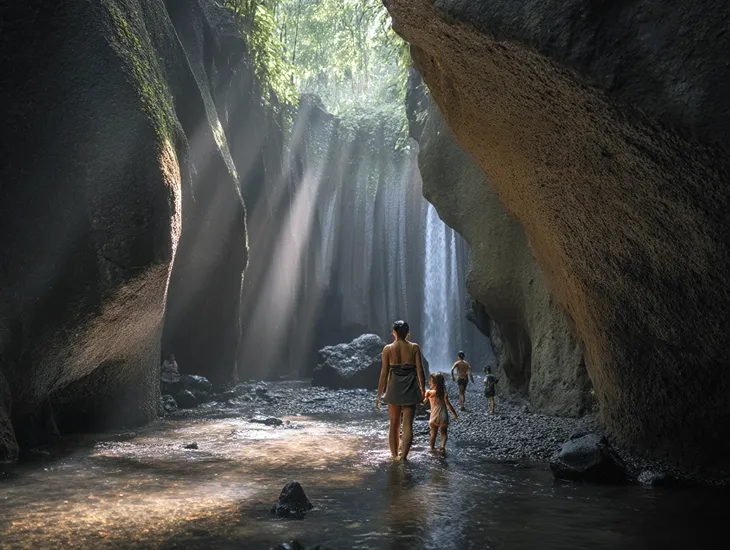 Tukad Cepung Waterfall in Bali – A caregiver guiding a child through a narrow rock passage toward the sunlight beams.