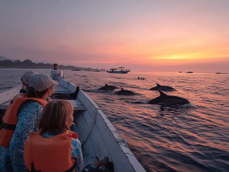 Bali Sunrise Tours 2026 – A traditional outrigger boat at dawn near Lovina with dolphins jumping in the distance before heading to Menjangan Island.