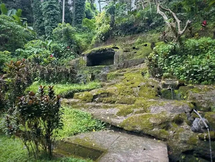 family adventure Goa Gajah in Bali 2026 – A child wearing a brightly colored batik sarong and a yellow sash standing near a stone statue.